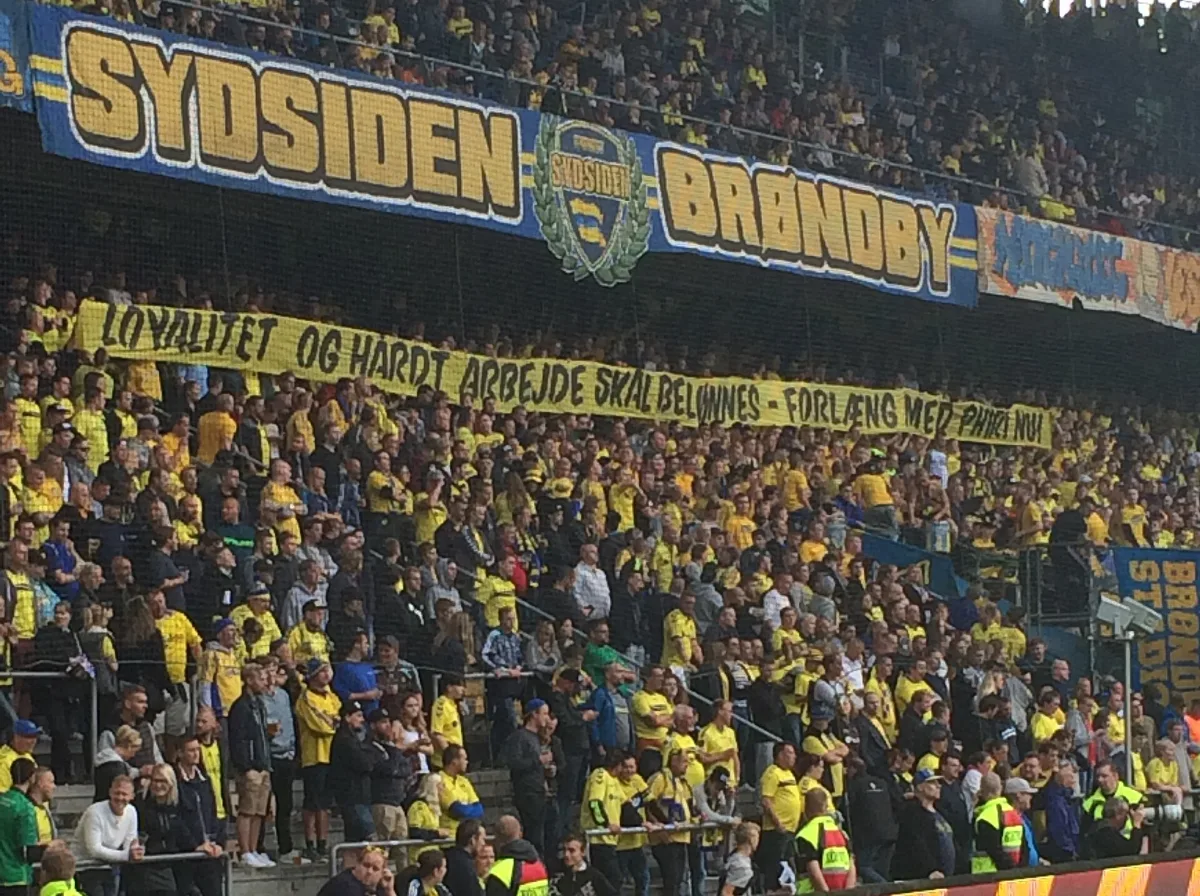The Sydsiden at Brøndby Stadion packed with yellow-clad supporters beneath a blue SYDSIDEN banner and a banner reading Loyalitet og hårdt arbejde skal belønnes – forlæng med Phiri nu!